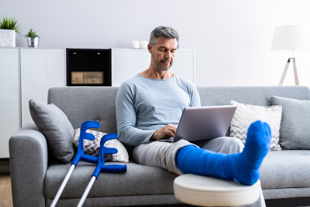 Eldre mann sitter i en sofa med brukket ben. Benet er i blå gips og hviler på en fotstøtte mens mannen har en laptop på fanget. To krykker står lent inntil sofaen