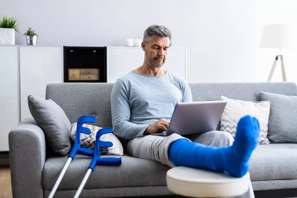 Eldre mann sitter i en sofa med brukket ben. Benet er i blå gips og hviler på en fotstøtte mens mannen har en laptop på fanget. To krykker står lent inntil sofaen